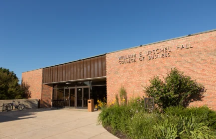 Brick building entrance with “William E. Urschel Hall College of Business” sign, plants, and clear sky—where the spirit of innovation shines like Beacon’s Reflection. Looking ahead to 2027, students like Esai Hernandez will continue to shape the future within these inspiring walls.