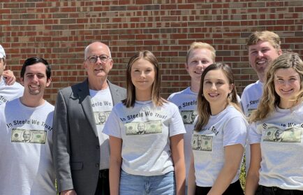 Group of people in matching “In Steele We Trust” shirts standing in front of a brick wall, smiling at the camera—members of Valparaiso University’s renowned Student-Run Investment Portfolio managing over $1 Million.
