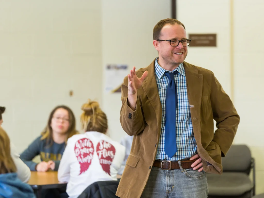 A man in glasses speaks to a group of students seated at tables in a bright classroom.