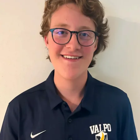 Smiling person with glasses wearing a navy "Valpo Business" polo shirt, representing Dean's Leadership, standing against a light wall.