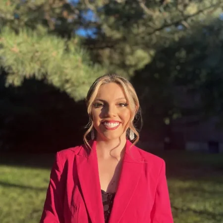 Woman in a bright pink blazer smiling outdoors, radiating confidence and Dean's Leadership against a backdrop of green trees and sunlight.