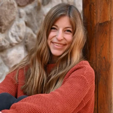 A woman with long hair in an orange sweater smiles while sitting by a wooden post and stone wall, exuding confidence and poise that reflect Dean's Leadership.