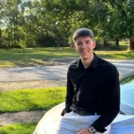 Young man in a black shirt sitting on a car, smiling confidently, embodying Dean’s Leadership, with trees and grass in the background.