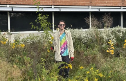 Valparaiso University environmental engineering student Alex Miroballi ’27 smiles as they hold up a long plant stalk in front of the Gellersen Engineering and Mathematics Center.