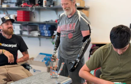 Three men are building robotics at a table; one man, a participant in the Beacon Academy Engineering Camp at Valparaiso University, has prosthetic arms and is smiling at the others.