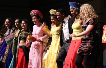 A diverse group in colorful cultural outfits stands together, smiling and linking arms on stage.