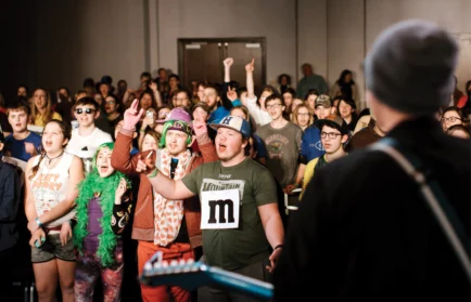 A performer on stage faces an enthusiastic crowd of young people singing along.