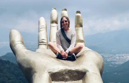 A woman sits cross-legged on a giant hand statue with mountains and sky in the background, embodying the spirit of Valparaiso University’s College of Nursing and Health Professions.