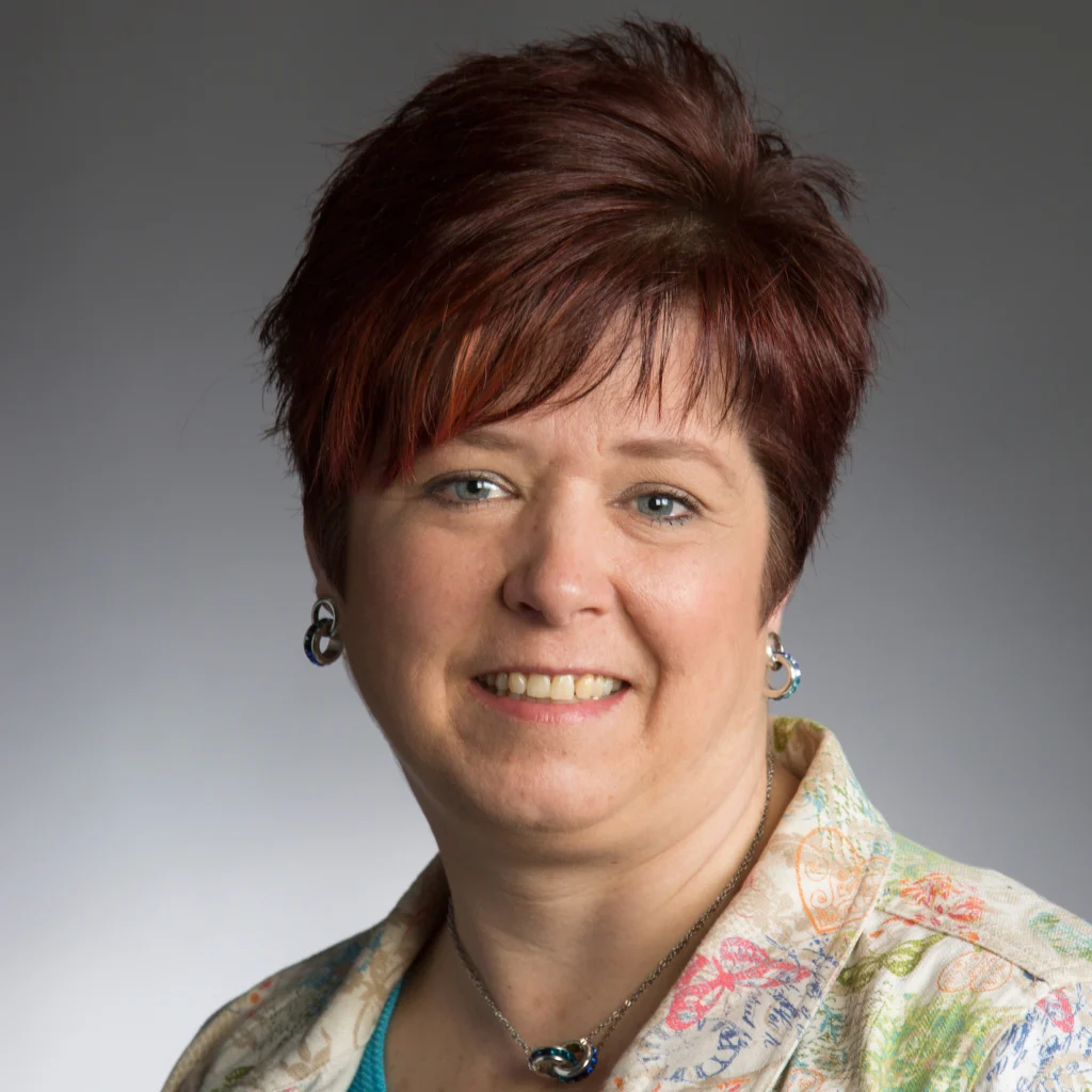 Smiling woman with short, reddish-brown hair, radiating academic leadership, wearing earrings, a necklace, and a patterned shirt.