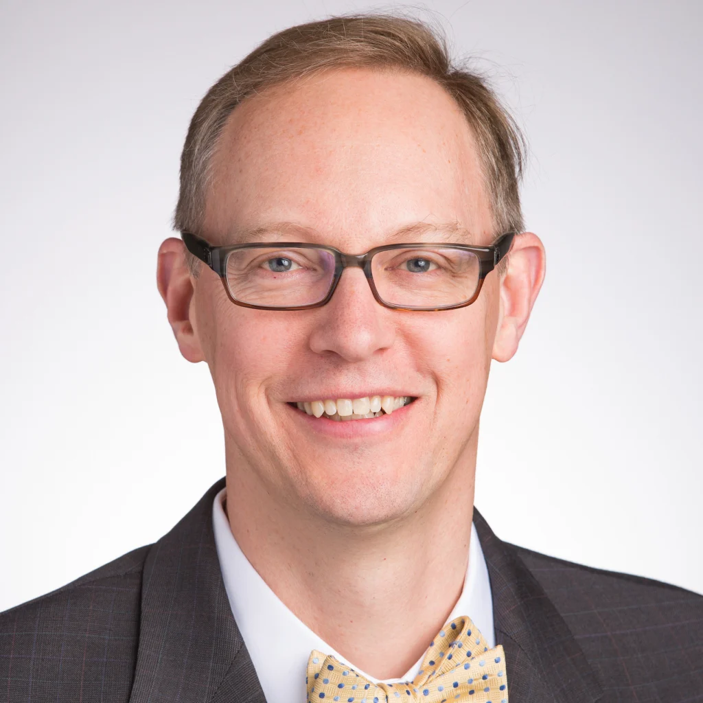 A man in glasses wearing a suit and bow tie, smiling at the camera against a light background, embodying academic leadership.