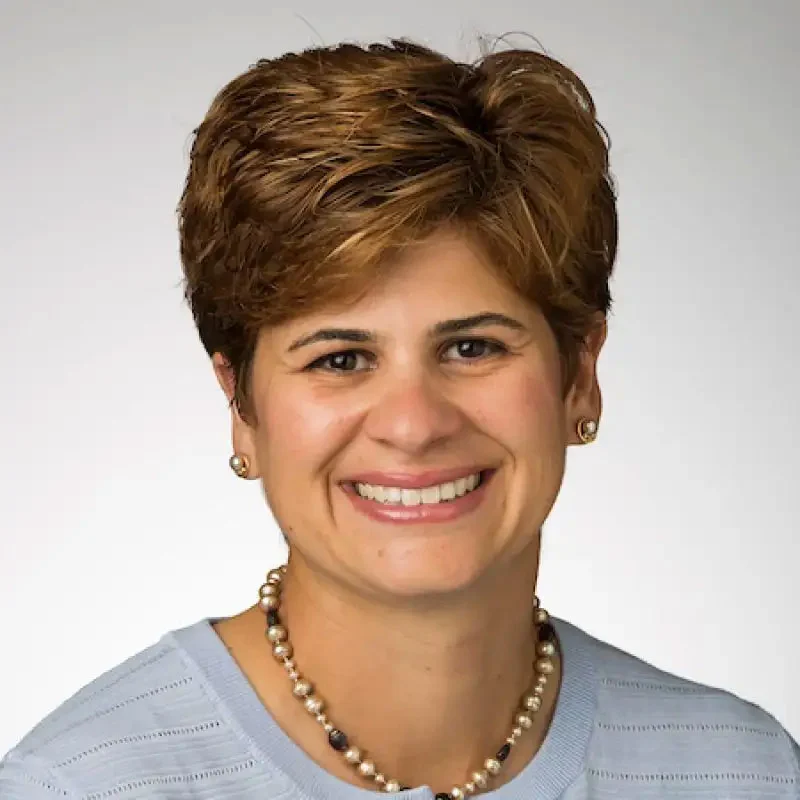 Smiling woman with short brown hair, wearing a light blue top, pearl necklace, and small earrings—radiating confidence and academic leadership.