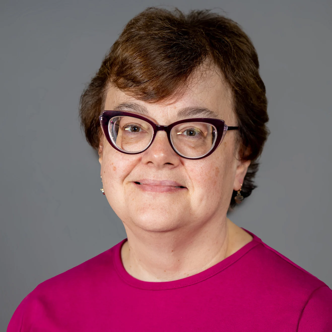 Woman with short brown hair, glasses, and a pink top smiling confidently in front of a plain gray background, embodying academic leadership.