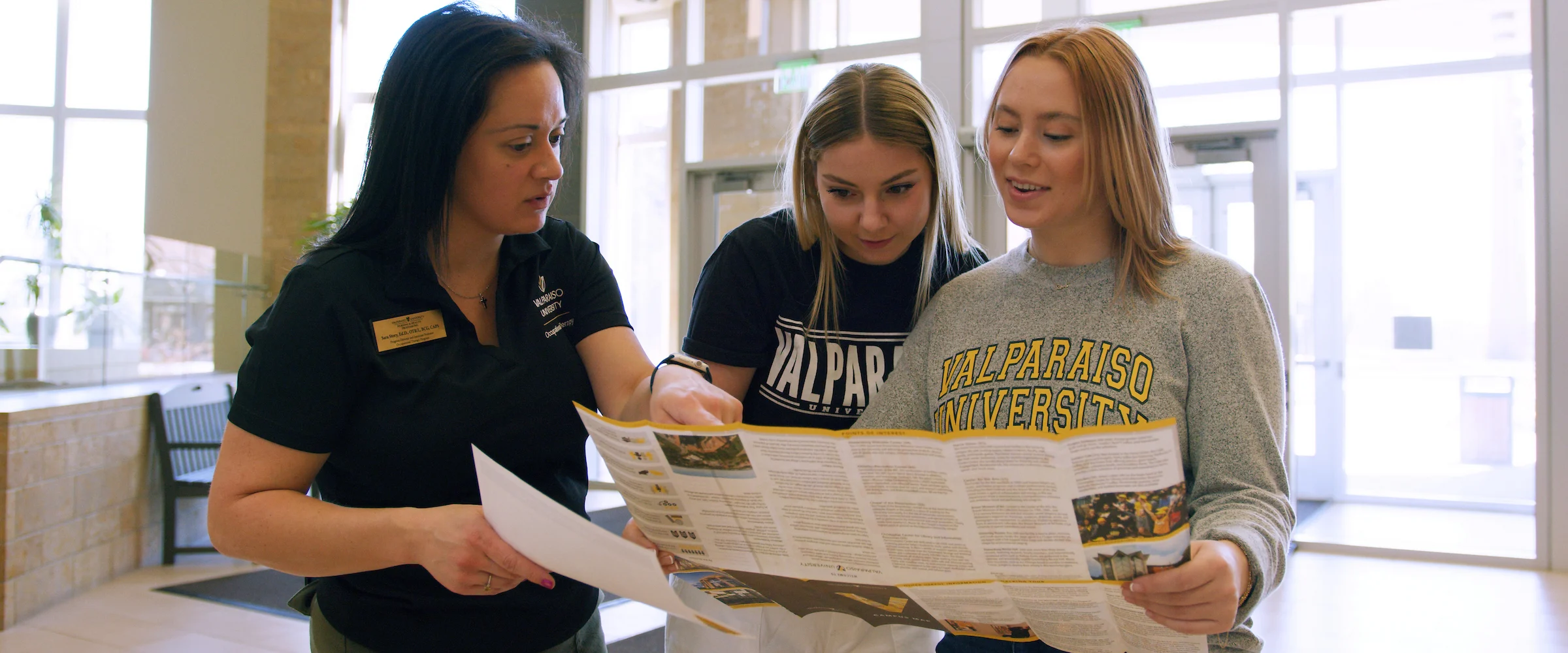 Three women look at a campus map together inside a brightly lit building with large windows, likely exploring options for Occupational Therapy Programs.