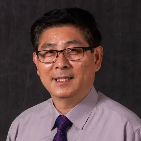 A man wearing glasses, a light purple shirt, and a tie, smiling in front of a dark background, exuding confidence and academic leadership.