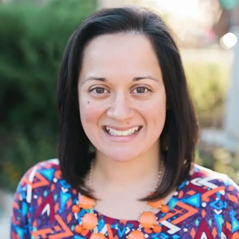 Woman with shoulder-length dark hair, wearing a colorful top and orange necklace, smiling outdoors, radiates confidence and academic leadership.