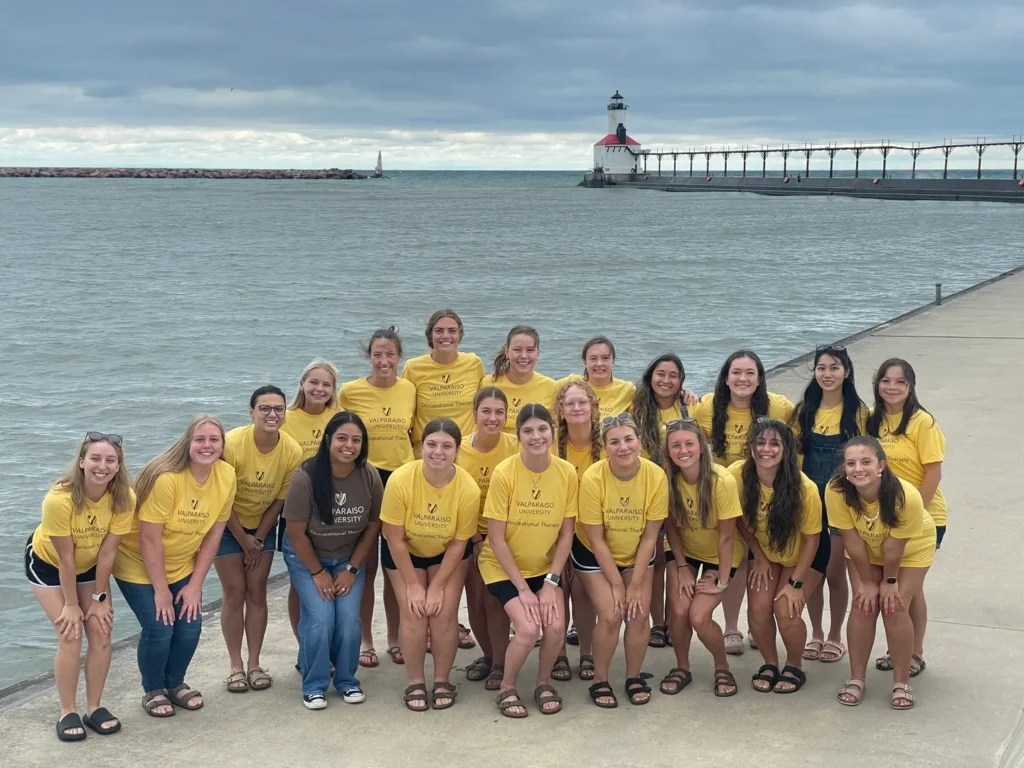 A group of women in yellow shirts, part of Occupational Therapy Programs, pose together by a lake with a lighthouse and pier in the background.