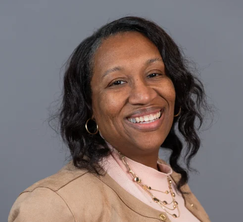 Smiling woman with wavy hair in a tan jacket and pink top, exuding confidence and academic leadership as she poses in front of a plain gray background.