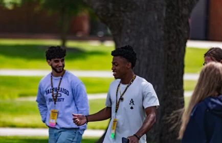 Two young men with name tags walk and talk outside, smiling, with trees and grass in the background, as they share college social tips on how to adjust socially during their first semester of college.