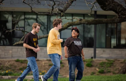 Three people walk together outside, talking and smiling, with trees and a modern building in the background—capturing the spirit of the Educating Character Initiative at Valparaiso University.