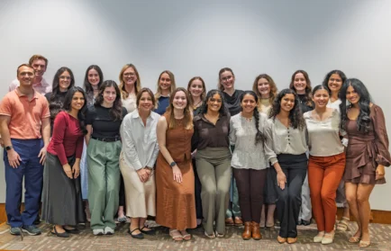 A group of 20 people smiling Valpo graduate studies students pose together in a well-lit indoor setting against a plain background, celebrating graduate Honors and their commitment to lead and serve.