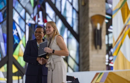 Two women smiling and posing with an award in front of colorful stained glass windows, celebrating a professional opportunity.