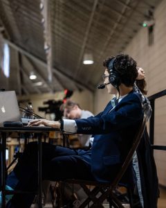 Dressed in a suit, Noah Godsell '24 looking away from the camera and towards the basketball court from WVUR-FM station, where he interacts with radio equipment to officially call the game.