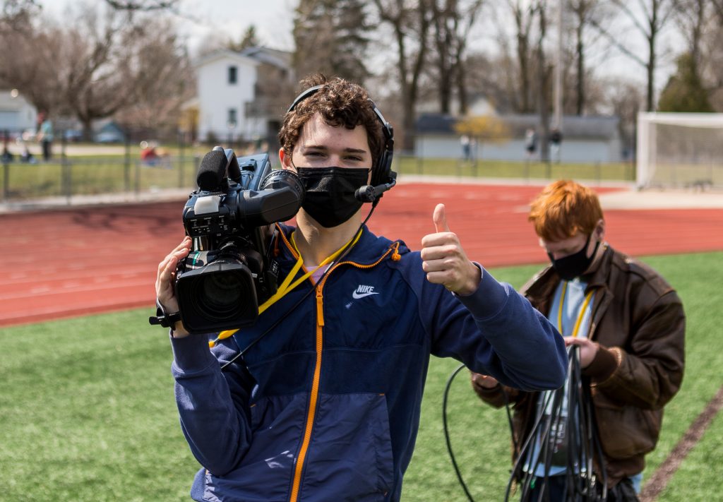 Noah Godsell '24 standing on the University soccer field, giving the camera a thumbs up with one hand and holding WVUR-FM filming equipment in the other, with the lower half of his face shielded by a protective mask.