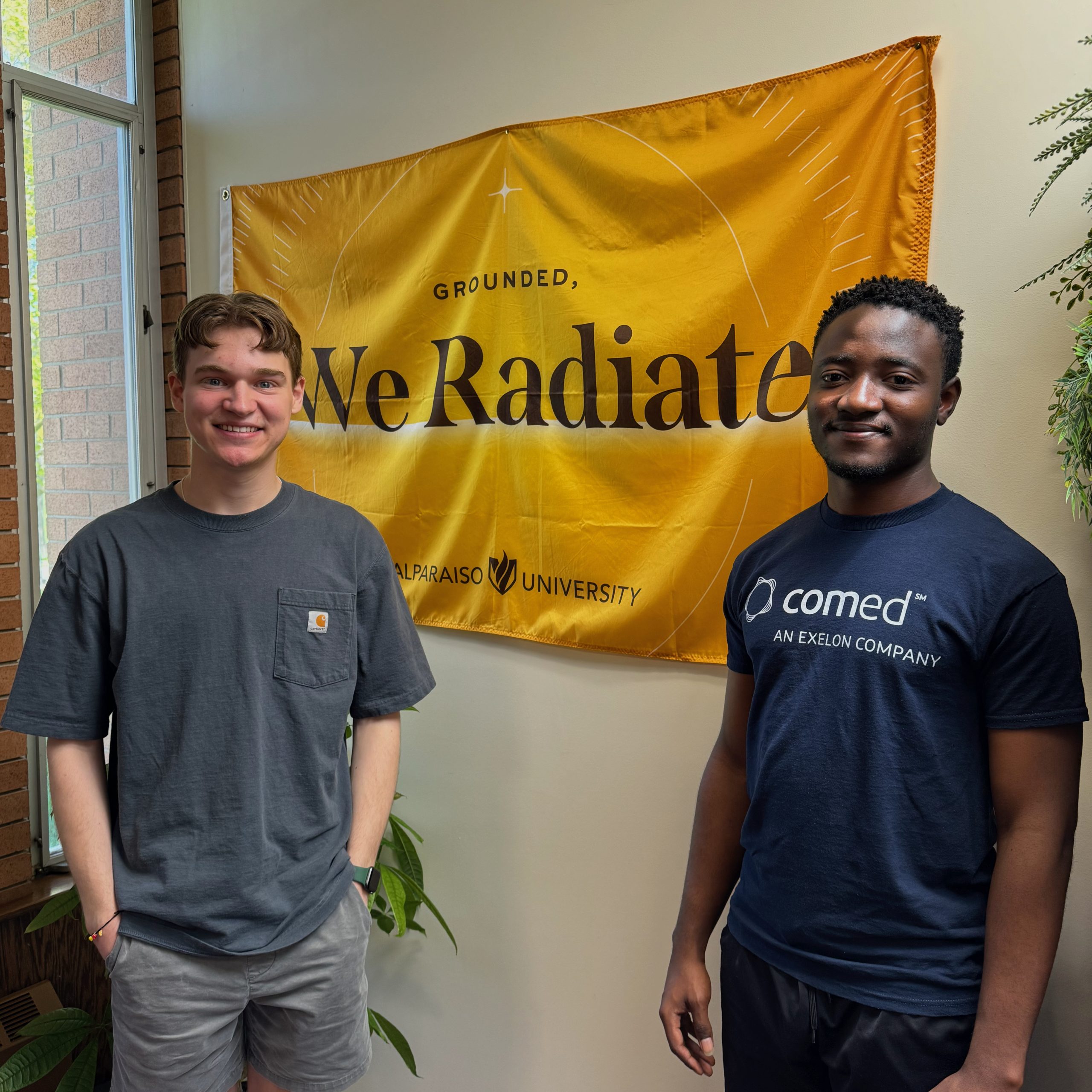 Charles Smith ’25 and Fayol Ateufack Zeudom ’26 smiling at the camera in front of a Valparaiso University flag reading, "Grounded, We Radiate".