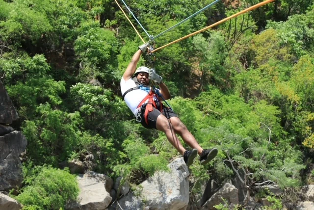 Antuane Thompson ziplines over a rocky, green landscape, wearing safety gear and a helmet—a true beacon of adventure and thrill.