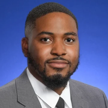 Antuane Thompson, dressed in a gray suit and tie, poses for a formal portrait against a blue background, serving as a beacon of professionalism.