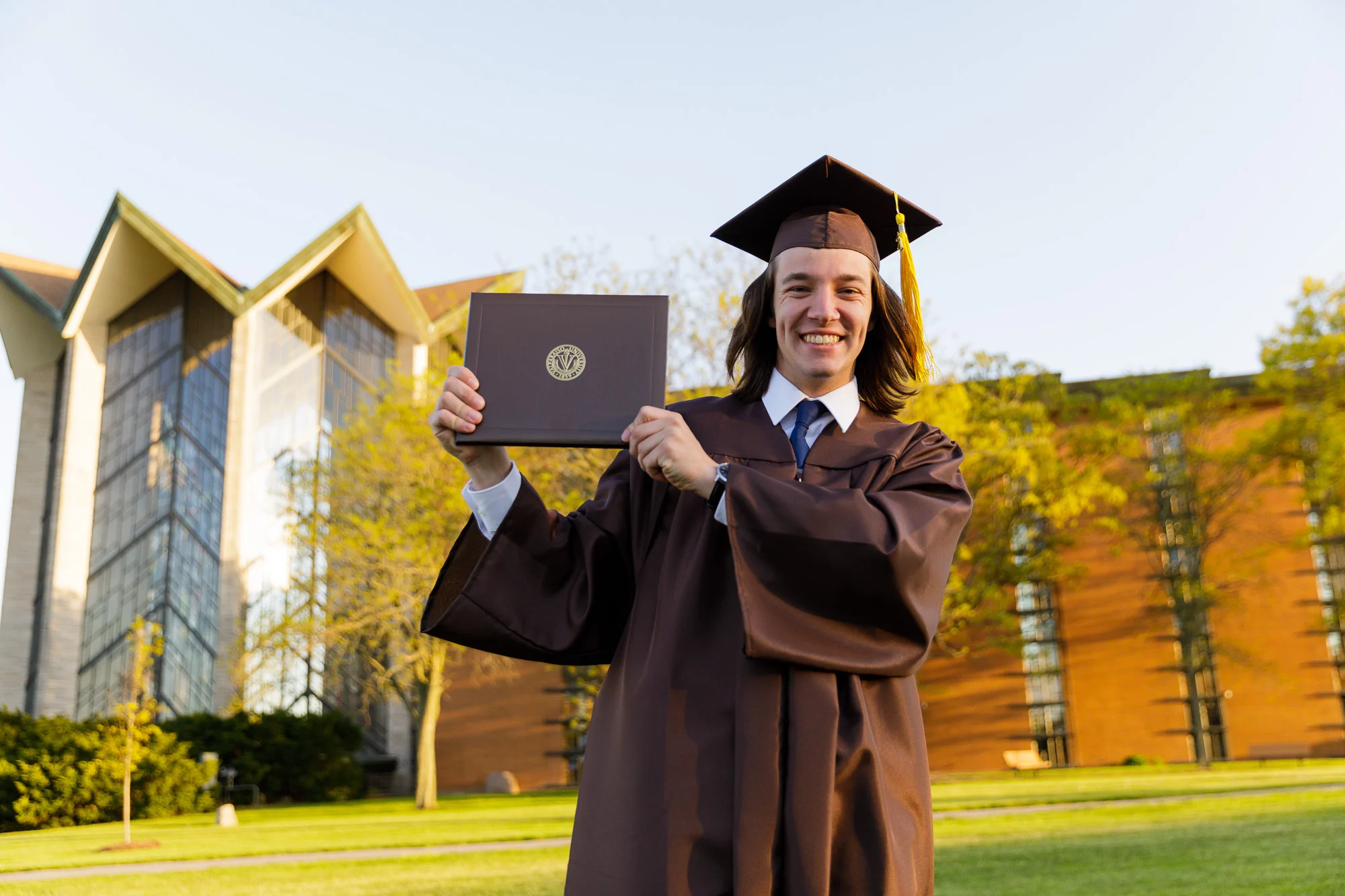 Smiling graduate in cap and gown holding diploma outside a modern building on a sunny day, discovering joy in every role—just like Andrew Benson.