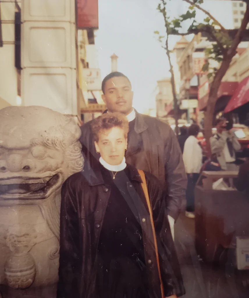 Two people stand in front of a stone lion statue on a busy city street, both wearing dark jackets. In the background, the word "Beacon" is faintly visible, adding intrigue to the scene.