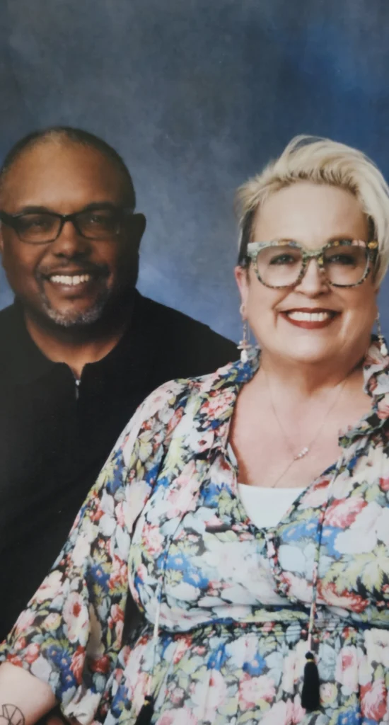 Thomas Barnett and Kim Wagner Barnett, both wearing glasses, smile together in front of a blue background, sharing a beacon of happiness.