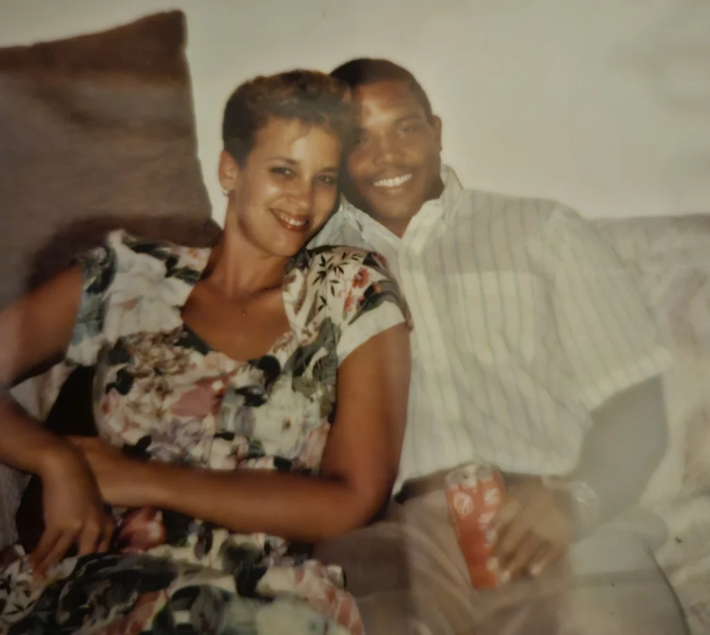 Smiling couple Thomas Barnett and Kim Wagner sit on a couch; she wears a floral dress, while he relaxes in a striped shirt holding a can—a cheerful Beacon moment.