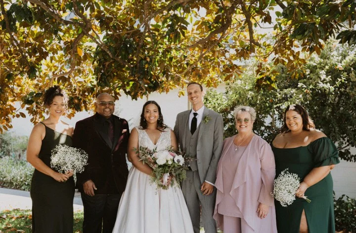 A wedding couple poses with family and friends under a tree, all smiling and dressed formally at the Beacon, celebrating Kim Wagner Barnett and Thomas Barnett’s special day.