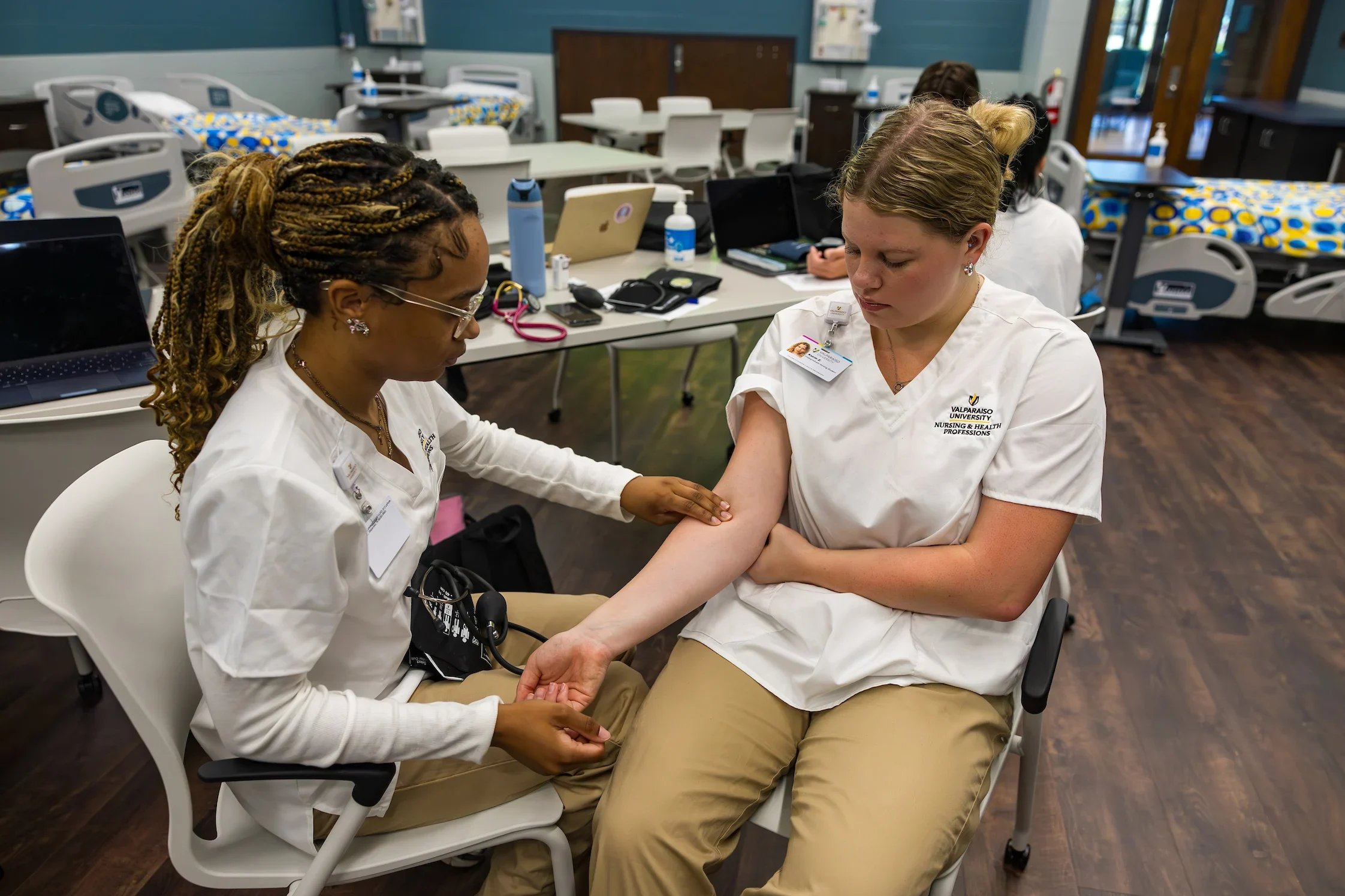 Two Valparaiso University nursing students practice checking a pulse in a nursing classroom equipped with medical beds and equipment.