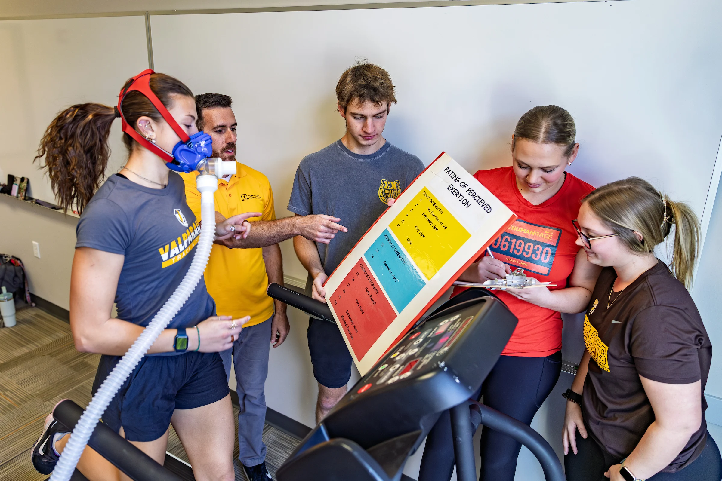 Three Valparaiso University students and a professor observe as a woman runs on a treadmill while wearing a breathing mask for a fitness test in an exercise science lab setting.