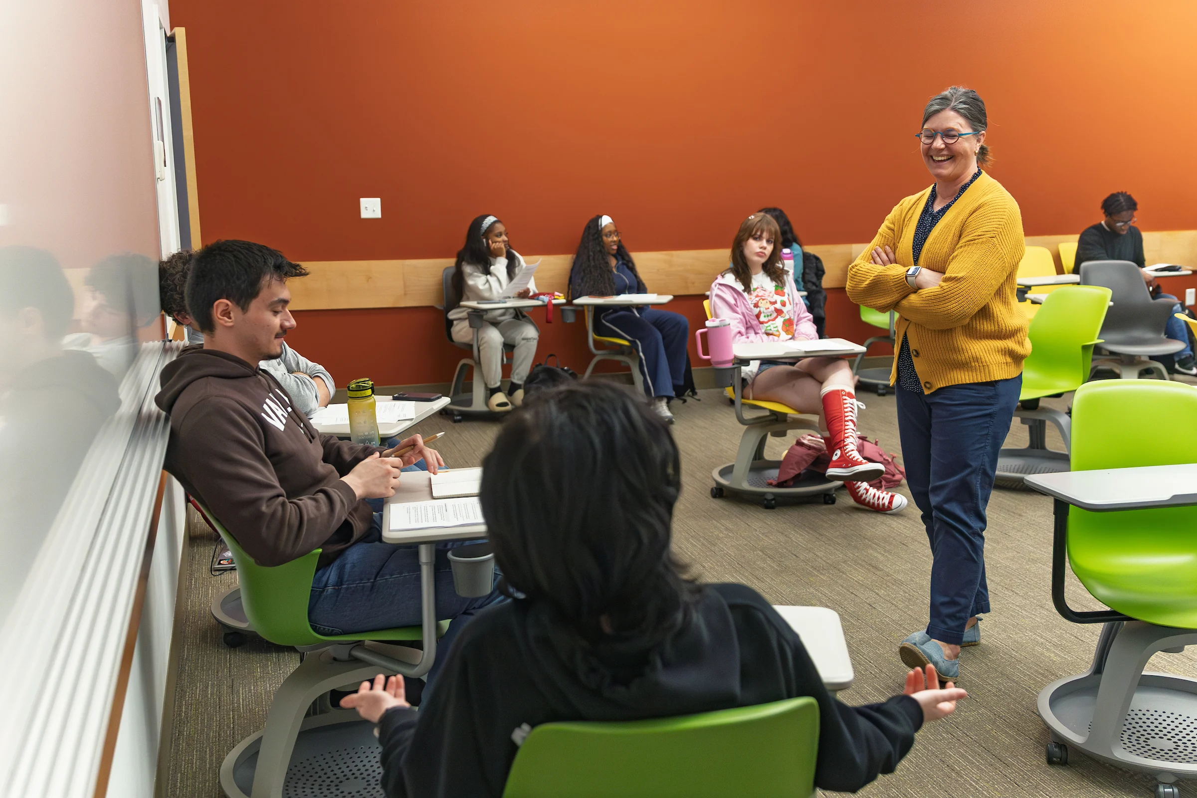 A teacher smiles while engaging with students seated in a colorful, modern classroom during a lively Religious Studies lesson.