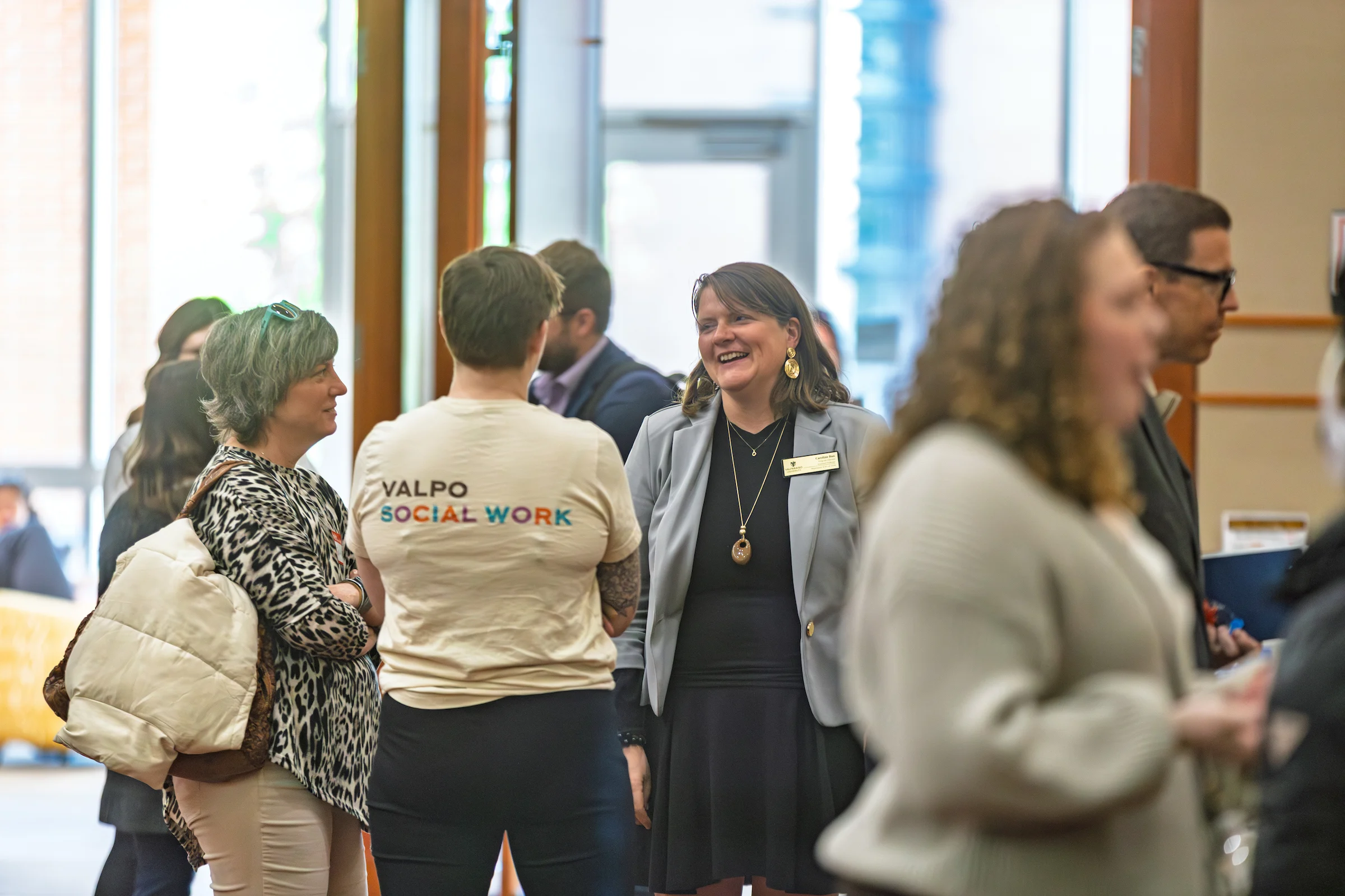 People gather and converse at an indoor event; one person, perhaps pursuing a Master of Social Work, wears a “VALPO SOCIAL WORK” t-shirt.