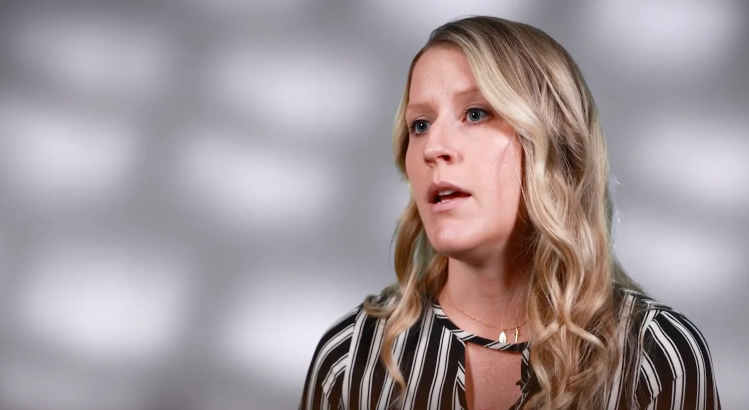 A woman with long blonde hair talks, wearing a striped top, against a gray background with light spots, sharing her insights from her MBA experience.