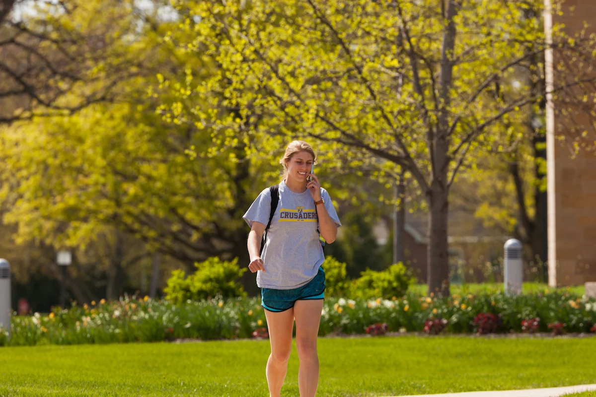 A young woman walks outside on a sunny day, talking on her phone about her upcoming commencement, with trees and grass in the background.