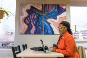 Woman in an orange jacket works on a laptop at a table with colorful abstract art behind her, perhaps researching the 10 Benefits of a Post-Professional Doctorate in Occupational Therapy.