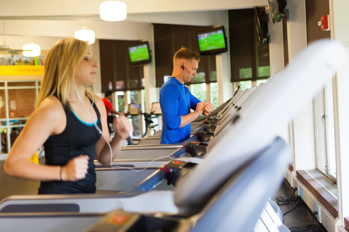 Two people focused on fitness run on treadmills in a gym, both sporting headphones and athletic clothing.