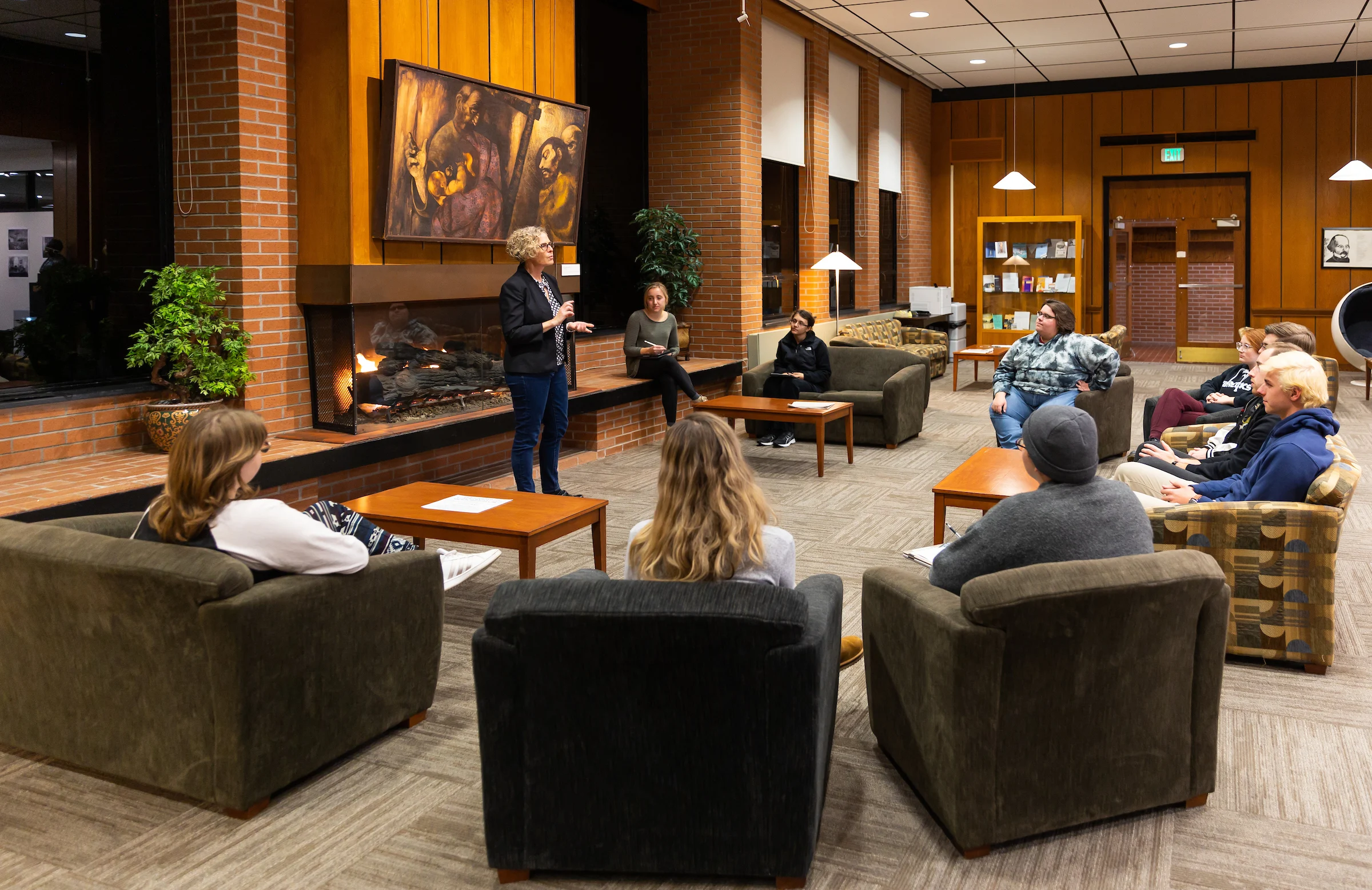 A group of people sit in a circle in a cozy room at Valparaiso University, listening to a speaker by the fireplace, engaging in faith-based learning.