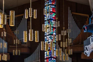 Gold cylindrical chandeliers and pipe organs surround a tall, colorful stained glass window inside a church, creating an inspiring atmosphere perfect for student involvement in community events and activities.