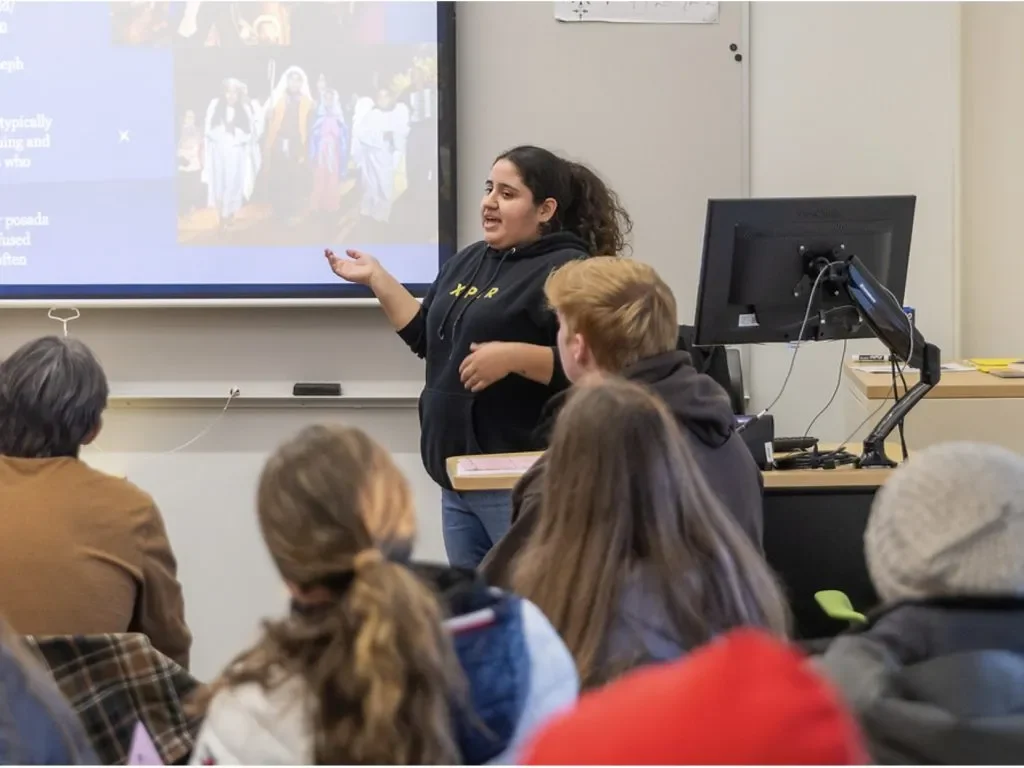 A woman gives a presentation to a classroom of students, gesturing toward a projection screen during a Faculty & Staff Groups meeting.