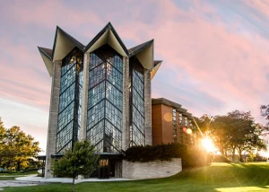 A modern glass Chapel of the Resurrection with an angular roof at sunset, surrounded by grass, trees, and soft pink clouds.