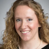 Smiling woman with curly blonde hair, wearing a black top and pearl necklace against a neutral background—Anne Marie Bice exudes effortless elegance.