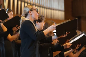 A group of people sings in a choir, holding black folders with sheet music in a warmly lit setting at a High School Choral Festival.