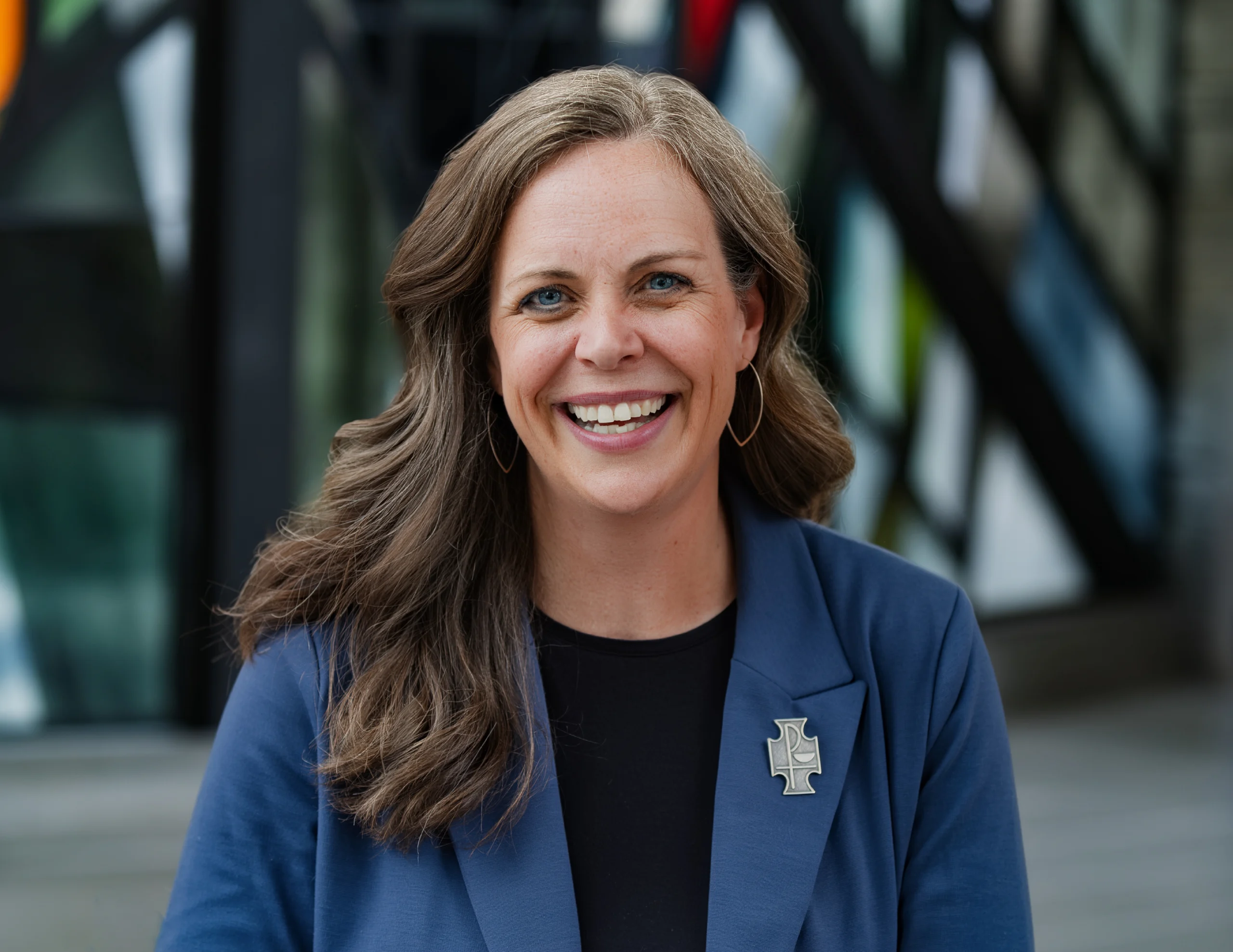 Kristin Lewis, M.A., with long brown hair and a blue blazer, smiles outdoors in front of abstract glass panels.